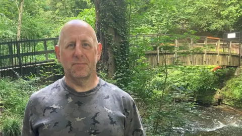 Photograph of Reece Sanford from Northern Moor in Wythenshawe in front of the Giant's Castle Bridge in Styal Woods. He is a bald man wearing a grey, black and white camouflage-style round-necked tshirt. The River Bollin flows under the wooden bridge.