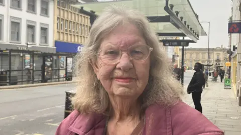 Mature woman wearing clear glasses and a mauve coat, smiling and looking directly at the camera. She has shoulder-length grey hair. She is standing near a bus stop with shops, a road and large buildings in the background.