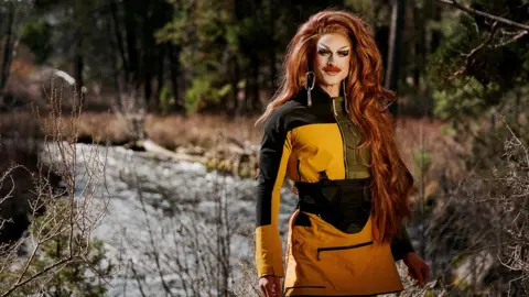 Performer Wyn Wylie, in drag as Pattie Gonia, wearing a yellow weatherproof dress with heavy face make up and long ginger hair. 