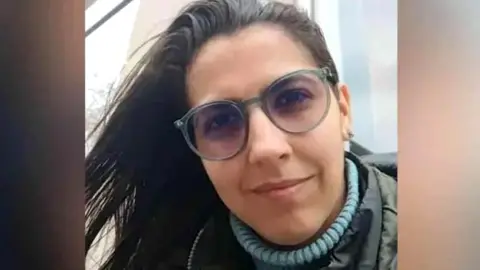 A picture shows the face of Barreto. She is wearing a blue turtleneck under a coat and glasses.