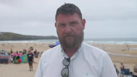 Mayor of Newquay Drew Creek stood on Fistral Beach. He is wearing a white shirt with black sun glasses. There are a number of people on the beach behind him. He is looking straight at the camera and has a big brown beard. 