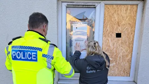 The back of a male police officer and female worker who is taping up a closure order on the window of the flat. The police officer has dark, short hair and is wearing a yellow hi-vis jacket with police written in white lettering on the back inside a blue box. The female worker has long, blonde, curly hair and is wearing a grey hoodie with white writing on the back which is covered by the hood. The closure order is on part of the window and the other side is covered up by wooden panelling.