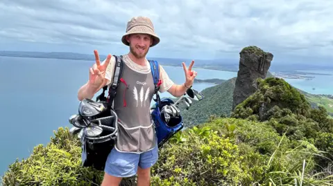 Dougie Haynes is doing a V peace symbol with both hands, whilst carrying two full golf bags on either shoulder. He is standing in front of a point where ocean meets land, with land visible on the other side of the water.