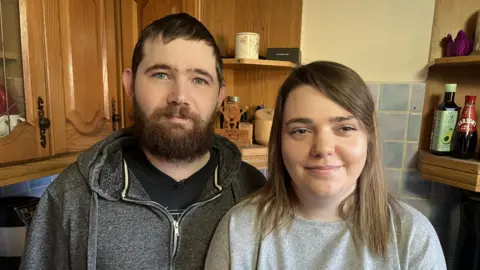 Abi is on the right, pictured with her partner on the left. Both are standing in their kitchen. Abi has shoulder-length brown hair and is wearing a grey jumper. Her husband has short brown hair and a beard. He is wearing a black t-shirt underneath a grey hooded zip-up jacket.