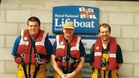 RNLI Three men in lifejackets and swim suits stand in front of an RNLI sign