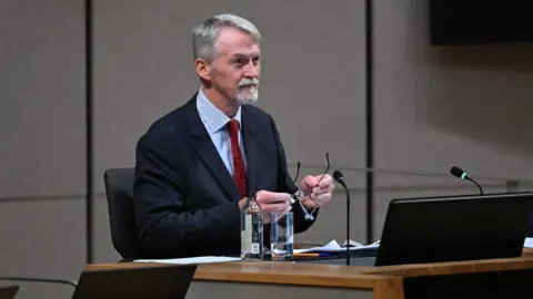 Senedd Cymru Huw Irranca-Davies in the Senedd chamber holding a pair of spectacles in his hands.