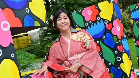 Pacemaker Women standing in red traditional outfit. Behind her is a background with different colours and green trees. 