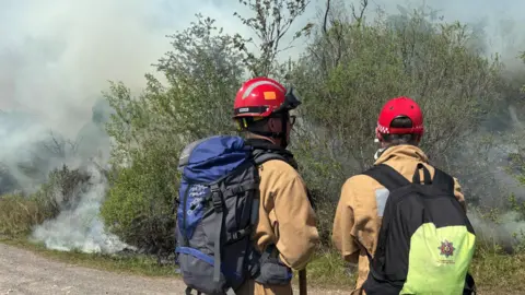 Two firefighters wearing large backpacks are looking at large plumes of smoke around gorse. They are wearing red hard helmets and tan fire resistant jackets.