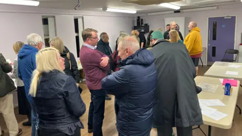 Residents and officials, all stood up and speaking with each other in a room, none of whom are looking into camera.