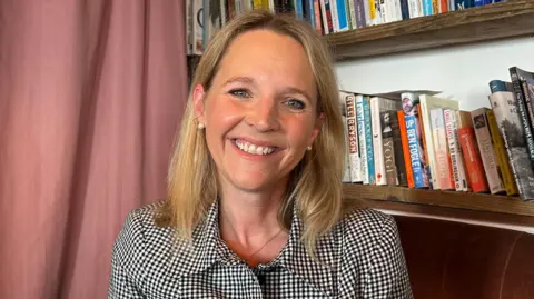 BBC Kate is seated on a brown upholstered chair in a cosy indoor setting, positioned in front of wooden bookshelves filled with a variety of books. She faces the camera and wears a short-sleeved, checked button-up shirt. A pink curtain hangs along the left side of the image, and soft indoor lighting illuminates the scene.