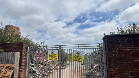 The bonfire site, which has been closed off by metal gates with signs reading "DANGER HAZARDOUS WASTE KEEP OUT", in the corner you can see the bonfire.