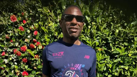 A man with sunglassed and a dark blue tshirt with purple writing is smilingat the camers as he stands against a bush with red roses on it