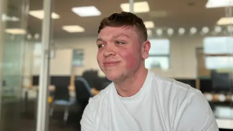 Lewis Miller at Lakes College. He has short, dark hair and wears a white T-shirt