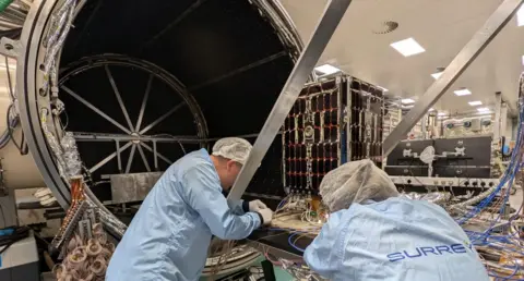 SSTL Jon Hall and Kai Berry from SSTL working on building a satellite. They are both wearing hair nets, rubber gloves and blue smocks. Various wires can be seen in the image alongside a partially constructed satellite.
