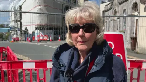 BBC The image shows a person standing in the foreground behind red and white temporary road barriers. The person is wearing a dark padded gilet over a long‑sleeved top and has a red lanyard around their neck. A small microphone is clipped to their clothing, suggesting they are being interviewed on camera.