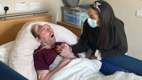 Alicia Manners A man lying in a bed with a tube running over his chest and into his nose. The man has a visible injury to his skull. A woman is stood at his bedside holding his hand. She has long dark hair and is wearing a white face mask. 