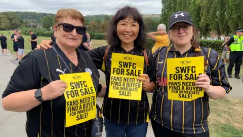 Three women are wearing black and yellow striped Sheffield Wednesday shirts. They are stood in a park holding yellow and black signs which say "SWFC for sale"