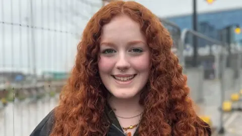 A young woman with long, wavy red hair looks into the camera. She is smiling and has a silver hooped nose ring.