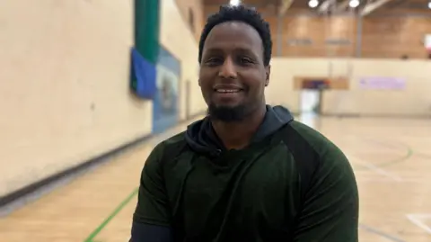 Abdi Jama sits in a sports hall. He has short dark hair and a short dark beard. He wears a green jumper