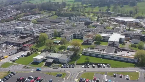 AIREDALE NHS FOUNDATION TRUST An aerial view of Airedale Hospital: car parks and green spaces can be seen in the foreground with an array of grey and white two-storey buildings behind. 