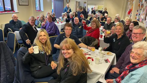 A group of men and women sat around tables in a community hall. They look into the camera, while some raise their glasses of orange juice.