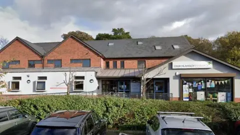 A view of a building housing a medical practice with a pharmacy attached to it. The building is mainly red brick with white rendering on the ground floor level. There are cars in a car park in front of the building
