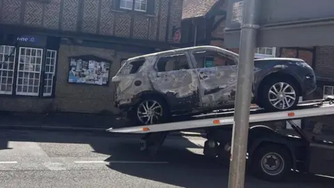 Nickita Starck A heavily damaged SUV is being transported on the back of a flatbed recovery truck. The bodywork is scorched and discoloured, particularly along the sides and rear, with missing paint and visible burn marks.
