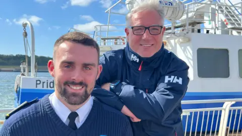 Head and shoulder shot of Stephen Jr and Stephen Snr - father and son. The younger man, to the left has a short dark hair, a close-cropped beard, and is wearing a dark blue v-neck jumper over a light blue shirt and dark tie. His father has grey hair, and is wearing spectacles and a navy blue jacket zipped up to the neck. Both are smiling and the Shields ferry can be seen just behind them.