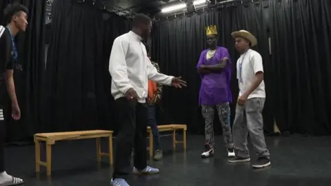 Five teenagers acting on a stage, one is wearing a straw hat and another a crown.