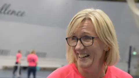 Jamie Niblock/BBC Helen is smiling and wearing glasses. She has a short, blonde bob, and is wearing a pink Suffolk Punch Pickleball club T-shirt.