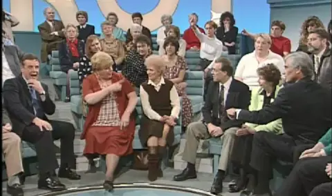 Haney in the audience of the TV show Kilroy. The host, a grey-haired man in a suit, sits to the right of the image. He appears to be trying to interject as Haney berates a woman to her left. All the audience are looking towards Haney. She is wearing a red skirt and jacket and is pointing at the woman next to her.