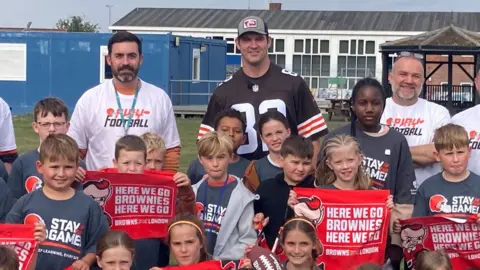 Um grupo de estudantes da Thrapston Primary School, vestindo James Buriz/BBC Gray T-Shirt, diz no jogo que vamos ao Brownies aqui com o Red Banner. Três homens atrás deles, uma camiseta branca de cabelos pretos e um professor com barba cinza e preta. O segundo é Gary Berniz, Sheven limpo, um boné de beisebol azul e verde claro e uma camiseta marrom; O terceiro é uma barba branca e outro professor do sexo masculino usando uma camiseta branca. Eles têm edifícios escolares atrás deles.