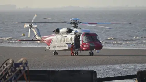 A red and white helicopter with the word "HM Coastguard" in black on the side. A crew member, wearing a yellow helmet and orange overalls, is standing on the sand next to the helicopter. Water is visible in the background.