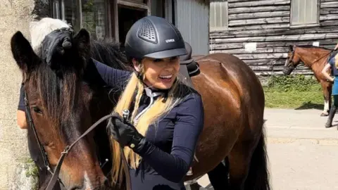 Ewa Larsson in dressage uniform, stood beside a brown horse in a yard. She is smiling and has a hand on the horse's mane.