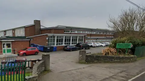 A school building. Several cars are parked outside the school. A sign outside says, Glastry College 