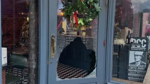 The glass panel on the front door, which is painted grey, has been smashed. A colourful Christmas wreath hangs on the door above the hole and the twinkling lights of the Christmas tree shine through from the shop inside.