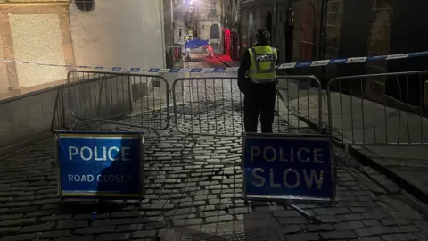 A police cordon on Niddry Street restricts access to Cowgate. The street is cobbled. On one side there is a sidewalk. On the other there is a wall. There are two blue signs on the road. Both have white writing. In one it reads 