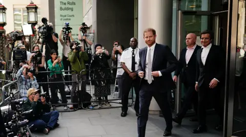 PA media Prince Harry settled out of court during his hacking case against the Daily Mirror. He wears a dark suit, white shirt and tie. To his left is barrister David Sherborne, also wearing a dark suit. A crowd of photographers is behind a metal barrier