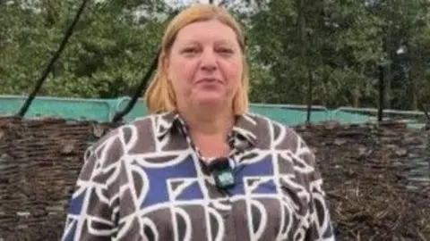 Jacqueline Mooney, with shoulder-length hair parted in the middle and wearing a geometric brown, blue and black shirt, stands in the centre of the charred roundhouse remains