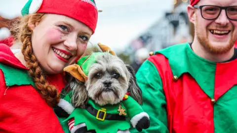 The Valley Evesham A woman with a long ginger plait smiles at the camera. She is wearing a red, green and white elf costume. A man on her right who is wearing an elf costume which is green and red with bells on is smiling too. In the centre they are holding a small dog which is also dressed as an elf, including a green hat with brown pointy ears. 