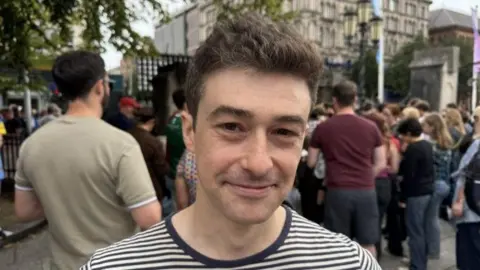 A man wearing a navy and white stripped t shirt is seen smiling at the camera as he stands in front of the city hall in Belfast ahead of the start of the Fleadh Cheoil na hÉireann with several dozen other people standing in the background.