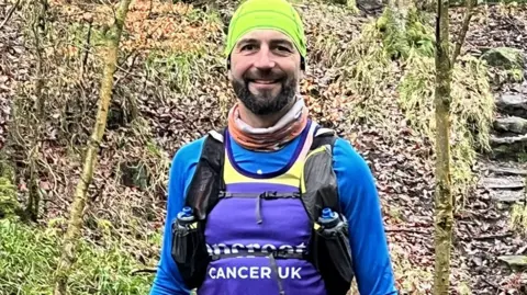 Oli Burbage-Hall A man smiles at the camera. He is wearing a bright yellow hat, an orange scarf, a long-sleeved blue T shirt and a purple vest. He is standing in woodlands.