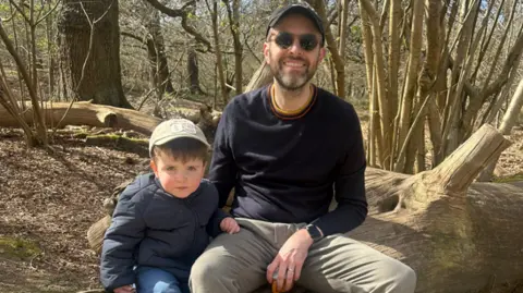 Dan Booth Dan is in a wood with Charlie. They both have caps on and Dan is wearing sunglasses. They are sat together on an old log.