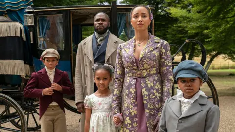 Liam Daniel/Netflix  The Mondrich family - an adult male and female with their three children, dressed in outfits from the 1800s, standing in front of a carriage looking up.