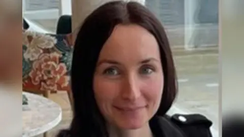 A woman with dark hair and blue eyes. She is smiling at the camera and wearing a black jacket.