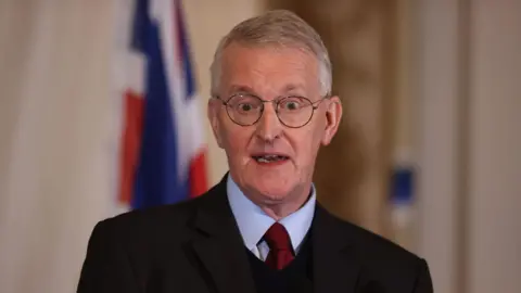 A grey haired man wearing round glasses looks to camera. He is wearing a navy suit, blue shirt and red tie.