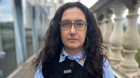 A woman with long dark hair looks at the camera. She is wearing green glasses.