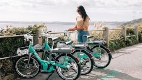 Dorset Council Three light green bikes standing up aside a path overlooking the coast, with a woman with sunglasses stood next to one, trying to get it started 