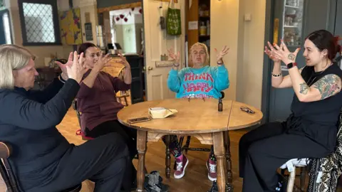 Anna Louise Claydon Participants sit around a table, all seated on wooden chairs, following the accessible warrior movement exercises