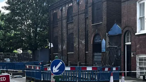 A two-storey brick building featuring arched windows with metal grilles and white graffiti. In the foreground, blue road barriers and a directional traffic sign surround a cordoned-off pavement area. A side entrance displays a Star of David.
